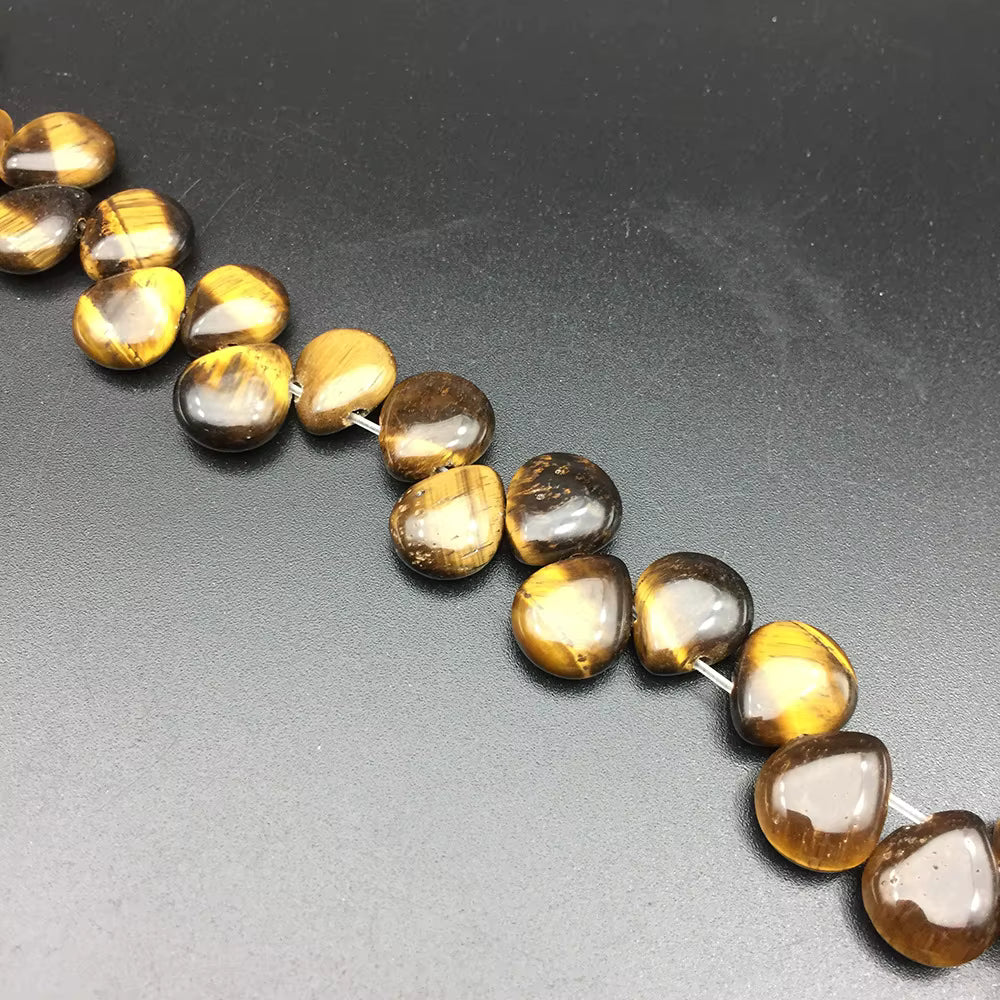 A close-up of a strand of polished tiger's eye beads on a dark gray background. The beads are teardrop-shaped with a glossy finish and display natural banding of golden-brown and dark brown.