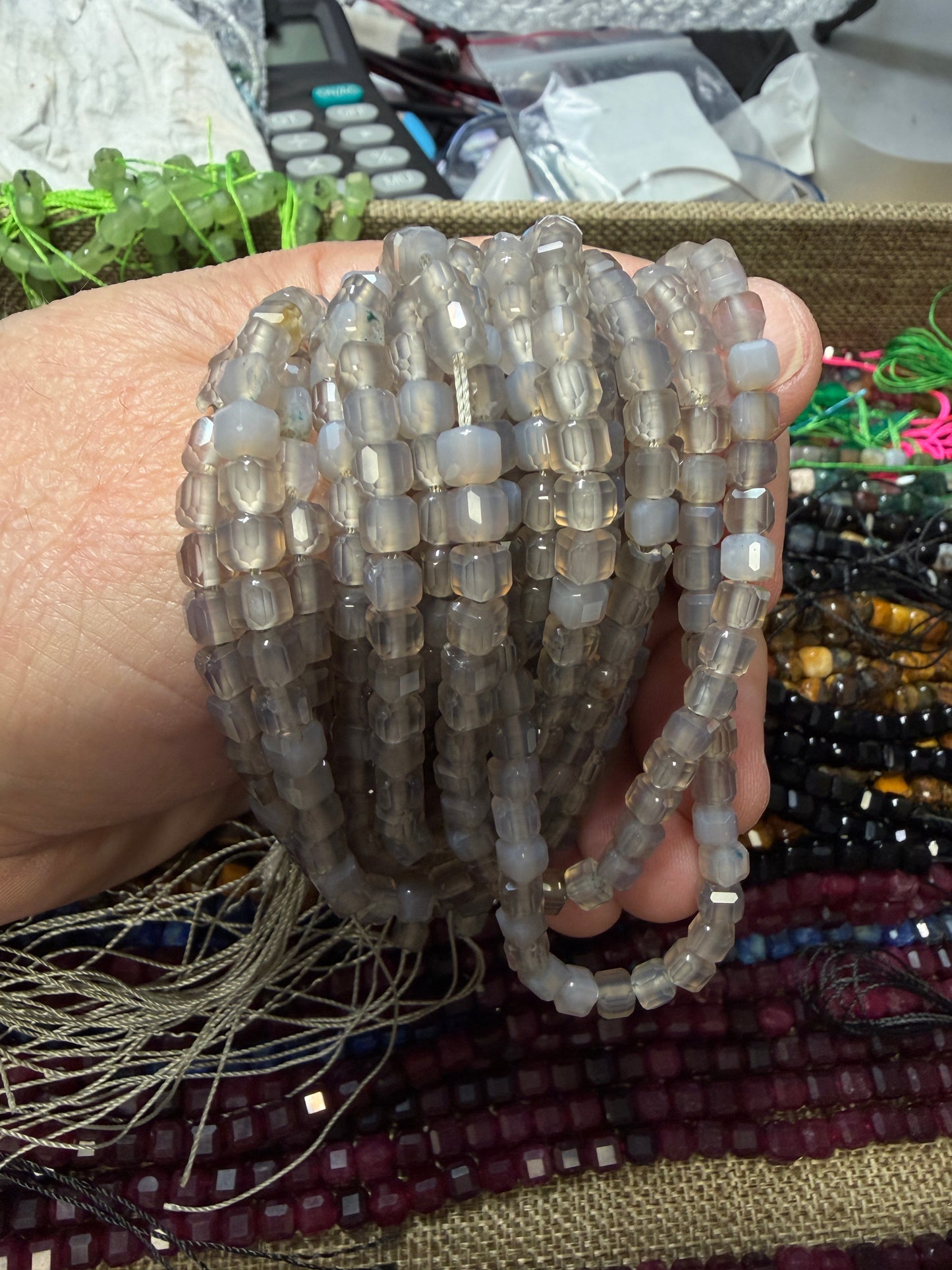 A hand holding a bundle of translucent gray faceted agate cube beads, with other beaded items and a messy desk background visible.
