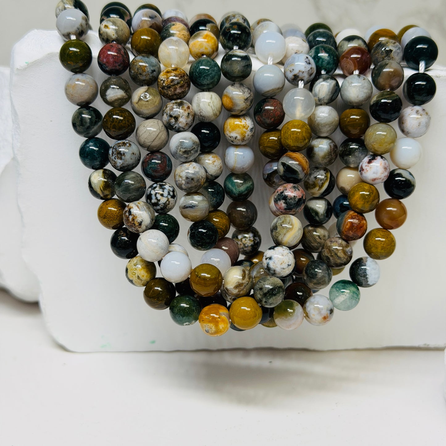 Multiple strands of polished round gemstone beads in assorted earthy colors and marbled patterns, draped over a white display block.
