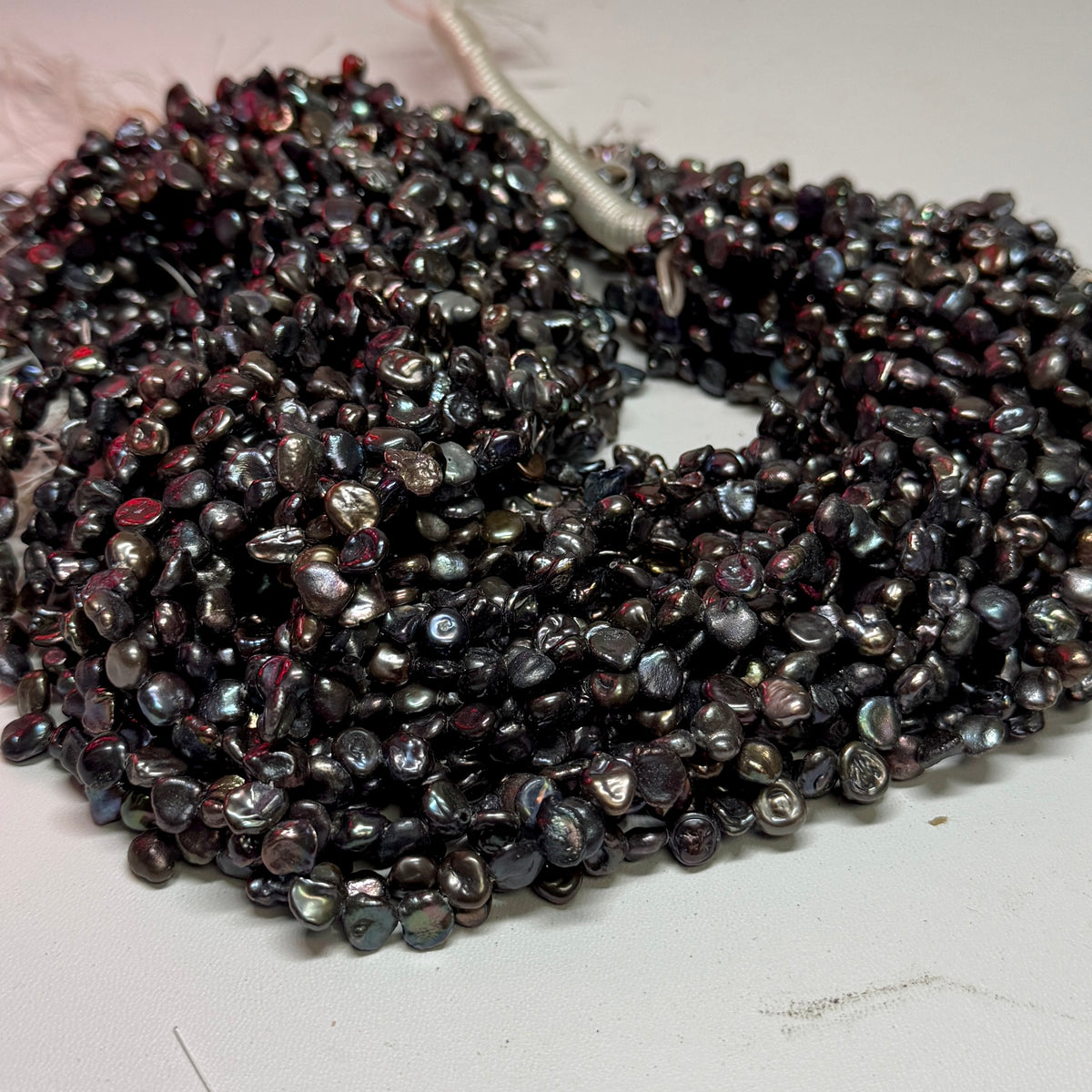 A pile of small, irregular keshi freshwater pearls with a dark, iridescent finish, showing hues of black, deep purple, and blue-green reflections, on a plain white surface.
