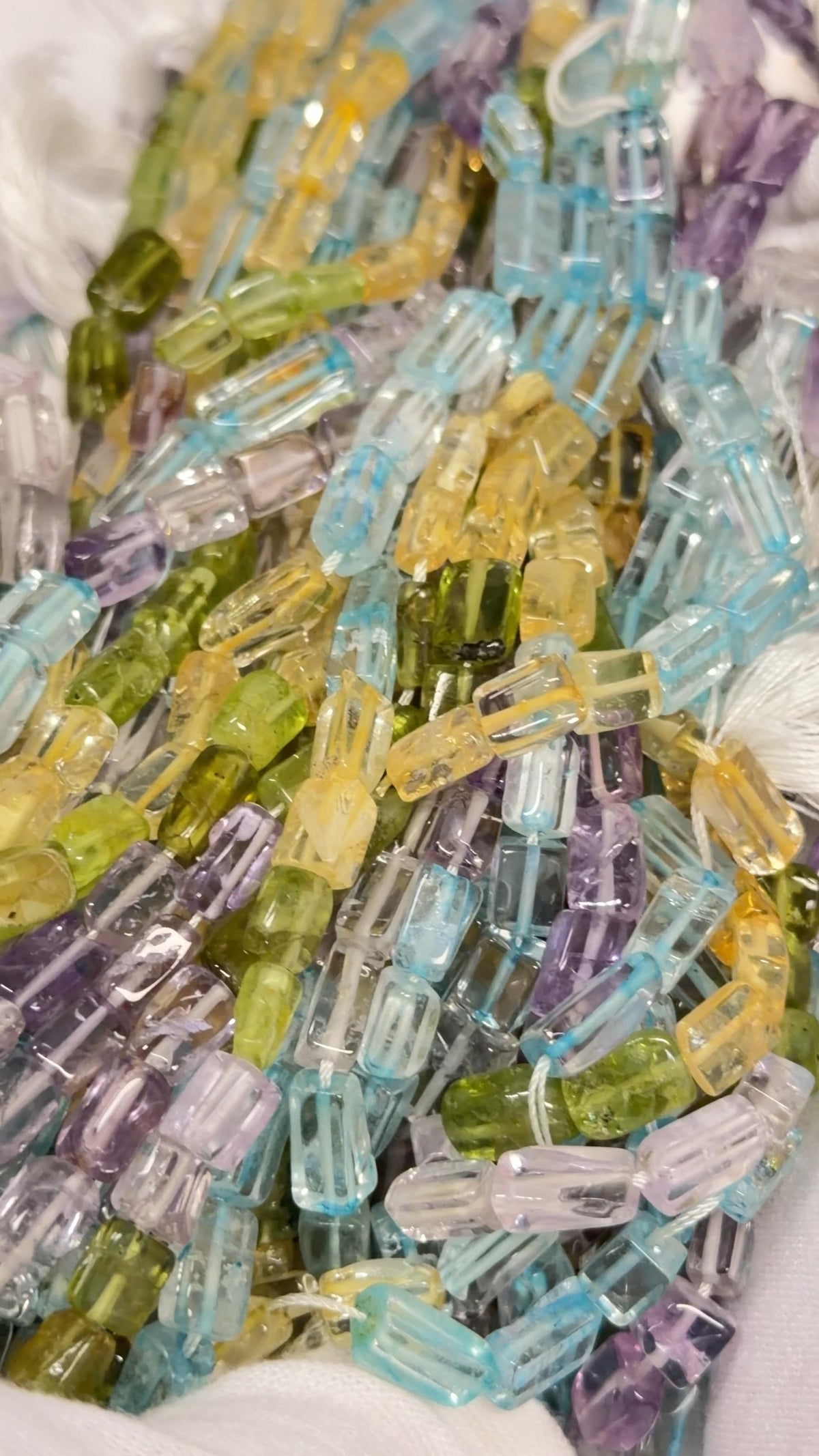 “Overlapping strands of translucent rectangular gemstone beads, showing rounded edges and purple, blue, yellow, green, and clear colors.”