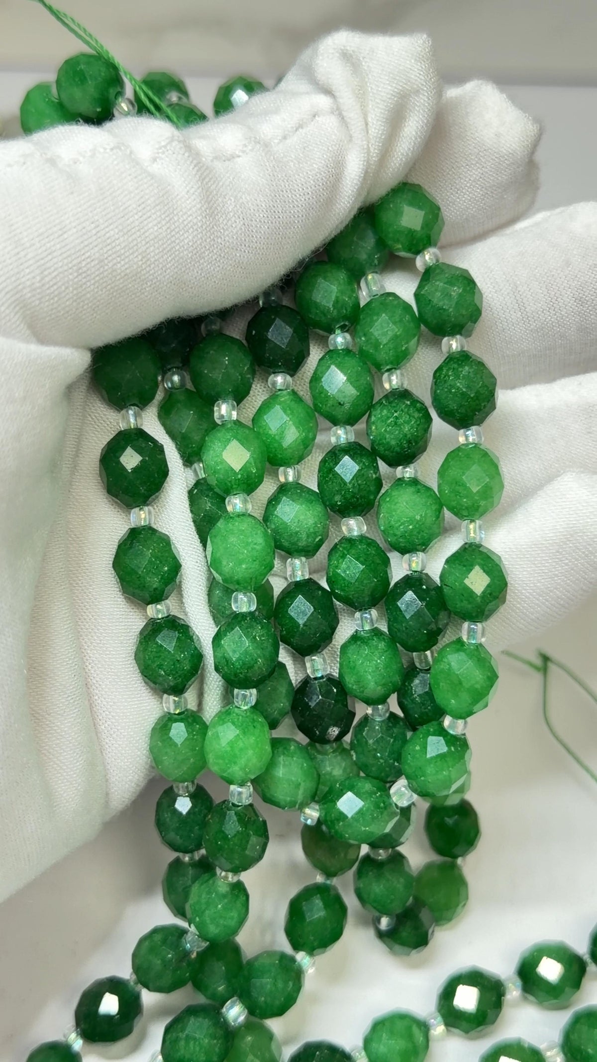 A close-up of a beaded strand made of round faceted beads in green tones, held in a white gloved hand.