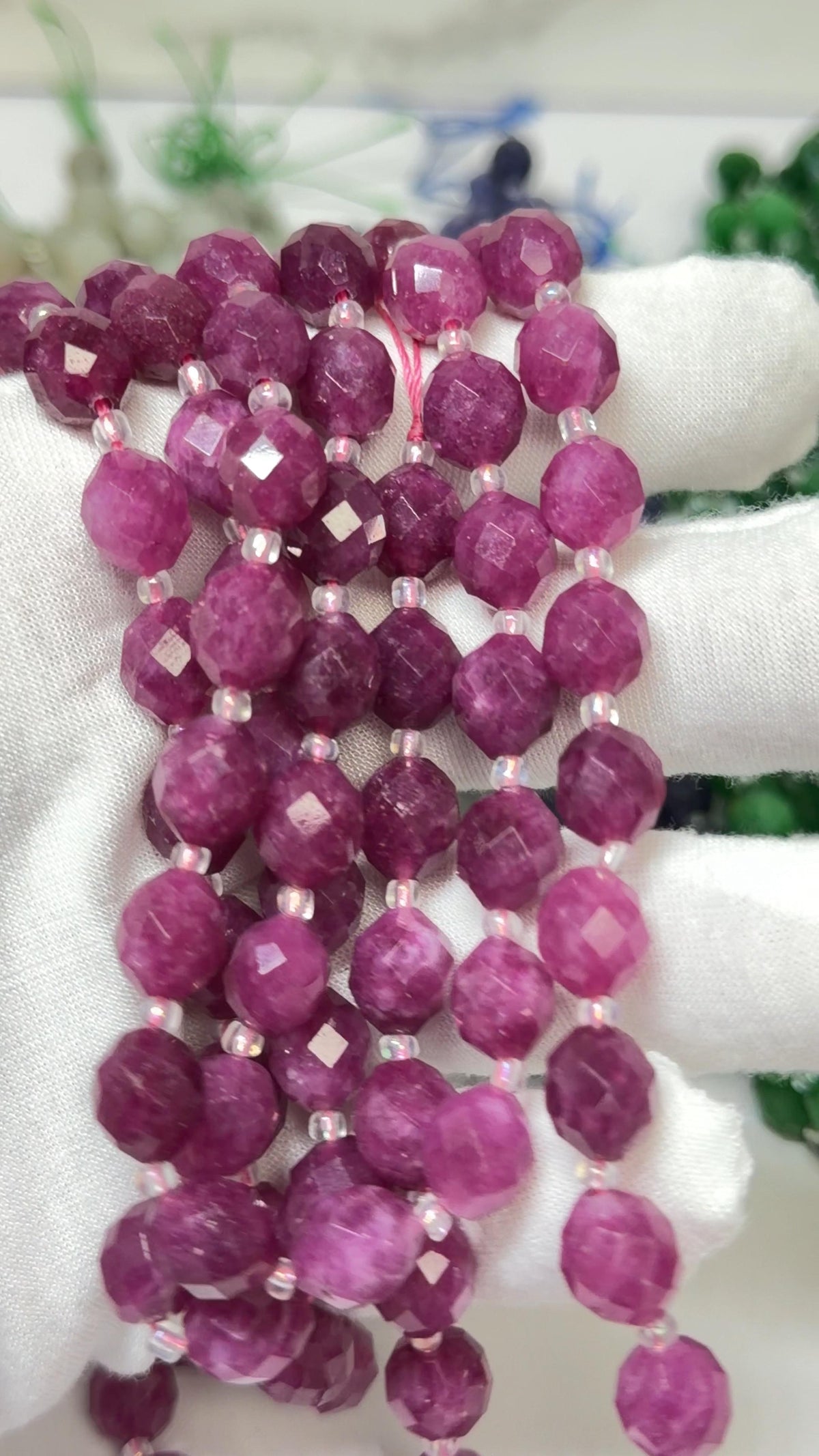 Faceted pinkish-purple gemstone beads strung on cords, held in a white gloved hand against a softly blurred background.
