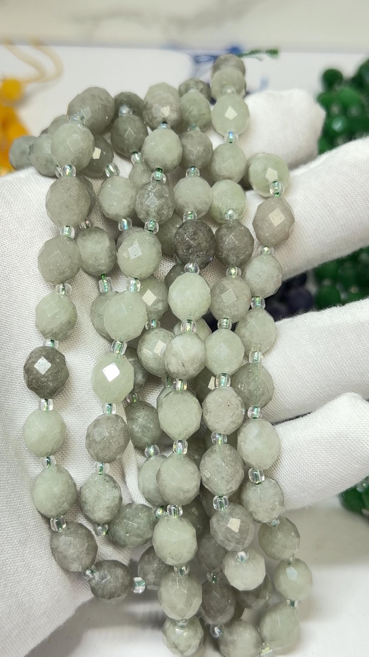 A close-up of a beaded strand made of round faceted beads in pale gray-green and taupe tones, held in a white gloved hand.