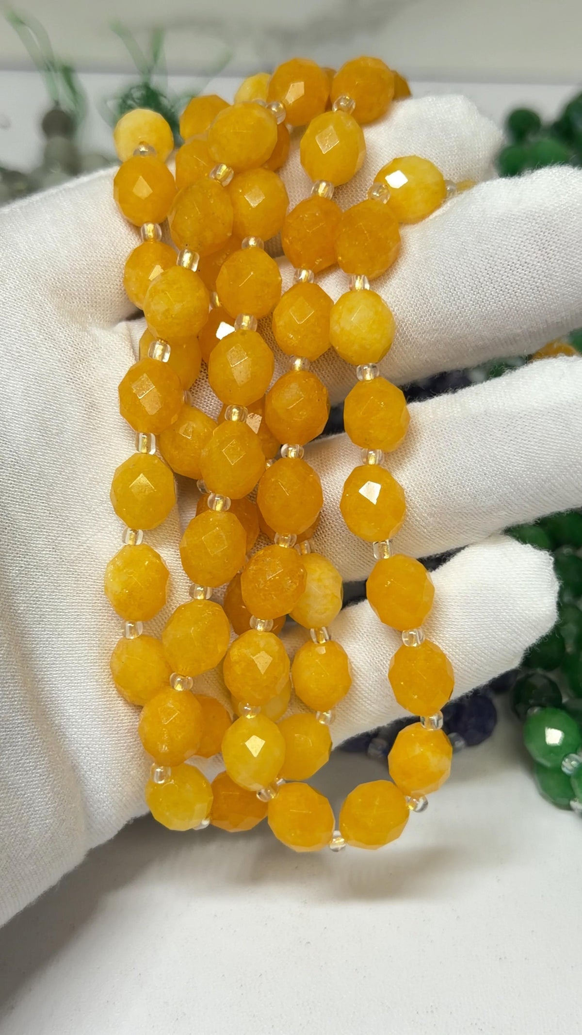 A close-up of a multi-strand yellow faceted bead necklace held in a white gloved hand, with other colored beads partially visible in the background.