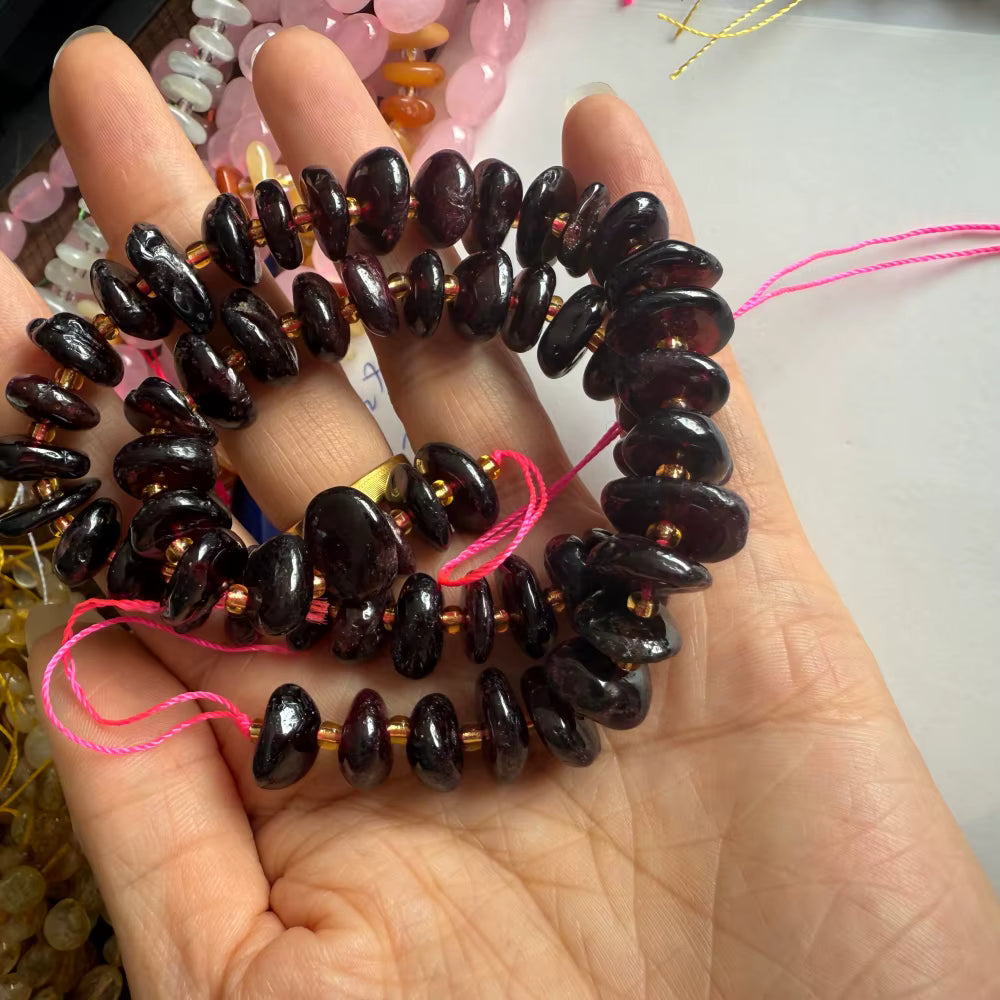 A hand holding a coiled strand of dark polished stone beads with small metallic spacer beads and pink thread ends, photographed against a light tabletop background.