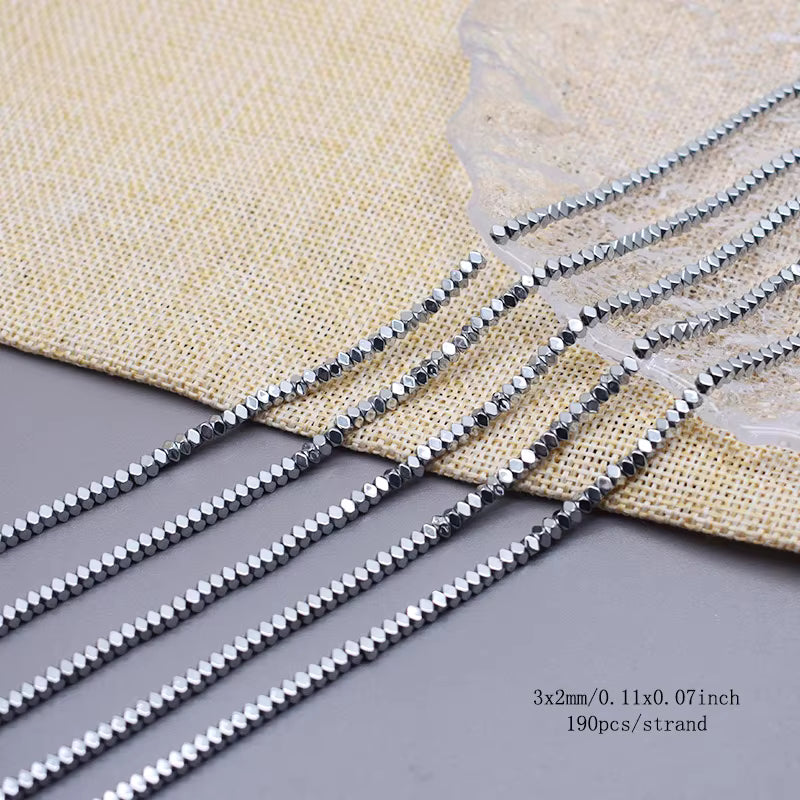 Five strands of diamond shaped hematite beads  on a gray surface with a beige woven fabric backdrop.  text in the bottom right specifies '3x2mm/0.