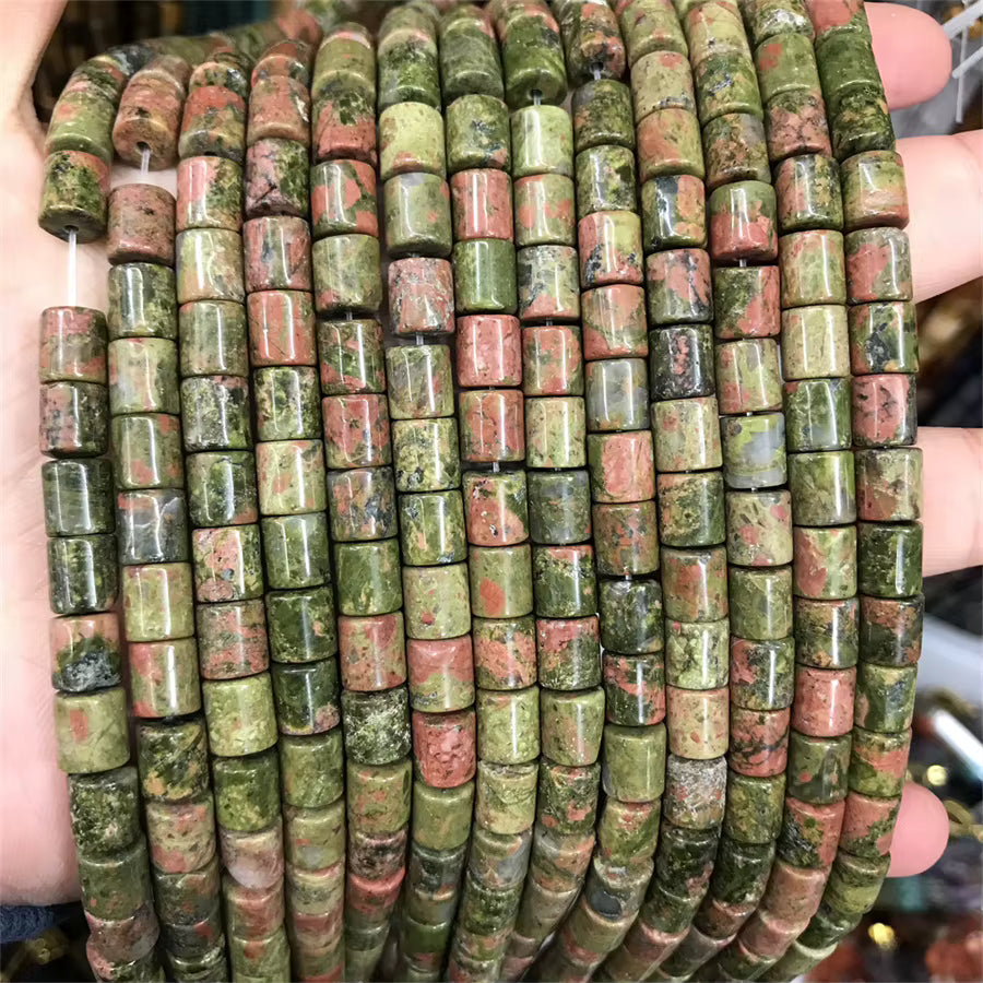 Multiple strands of cylindrical stone beads held in a hand. The beads have a natural marbled pattern in olive green, terracotta red, and dark brown, resembling jasper or rose quartz.