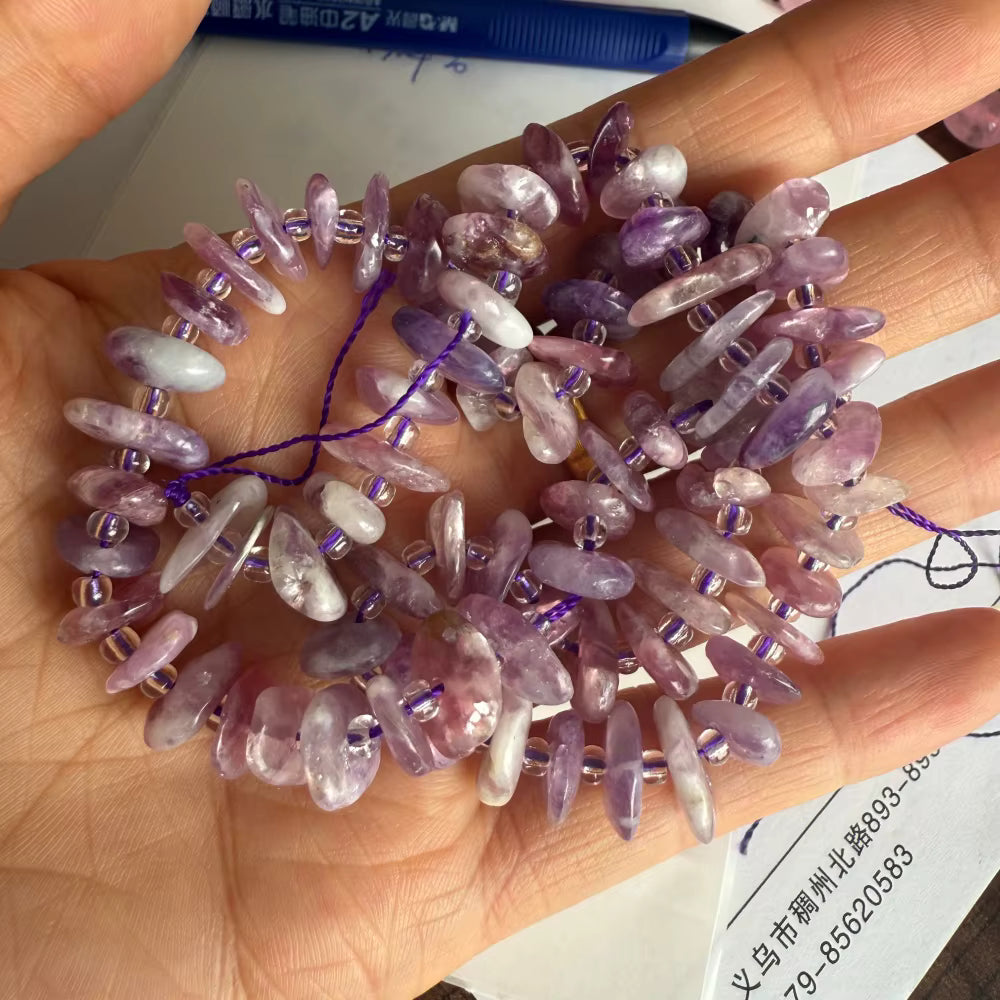 A hand holding a strand of irregular purple and pink amethyst beads with small clear spacer beads and a purple thread.