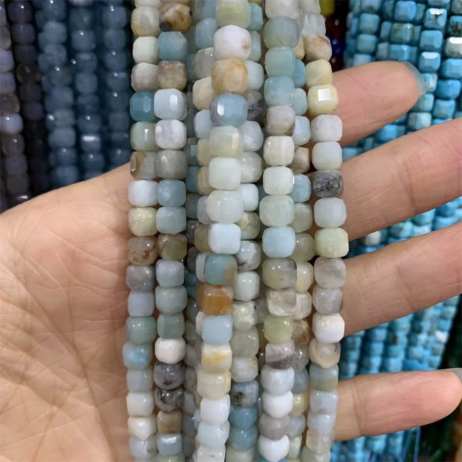 A hand holding several strands of faceted square beads in light blue, gray, white, and beige stone, with blurred blue beads in the background.