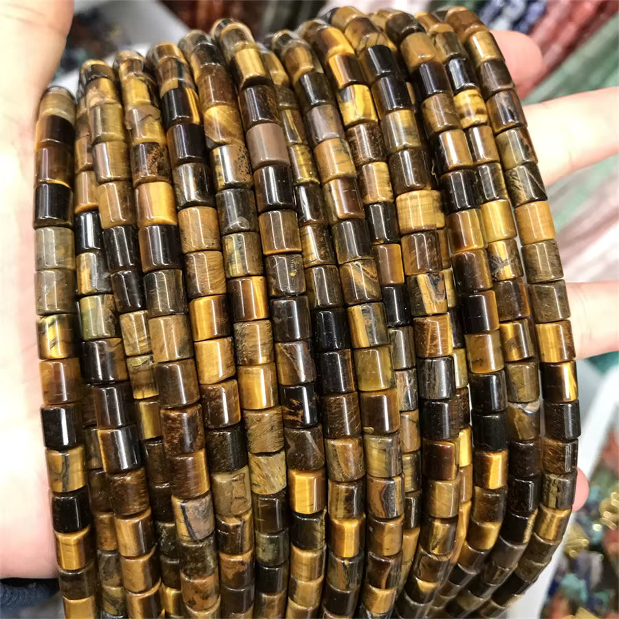 A close-up of multiple strands of cylindrical beads made from polished tiger's eye, held in a person's hands. The beads display natural banding with colors of golden yellow, brown, and dark brown to black.