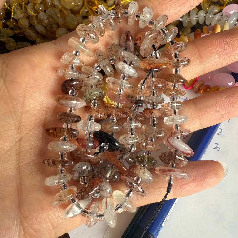 A hand holding a strand of polished translucent gemstone chips, likely quartz or amethyst, with mixed clear, smoky, and brown tones separated by small silver spacer beads.