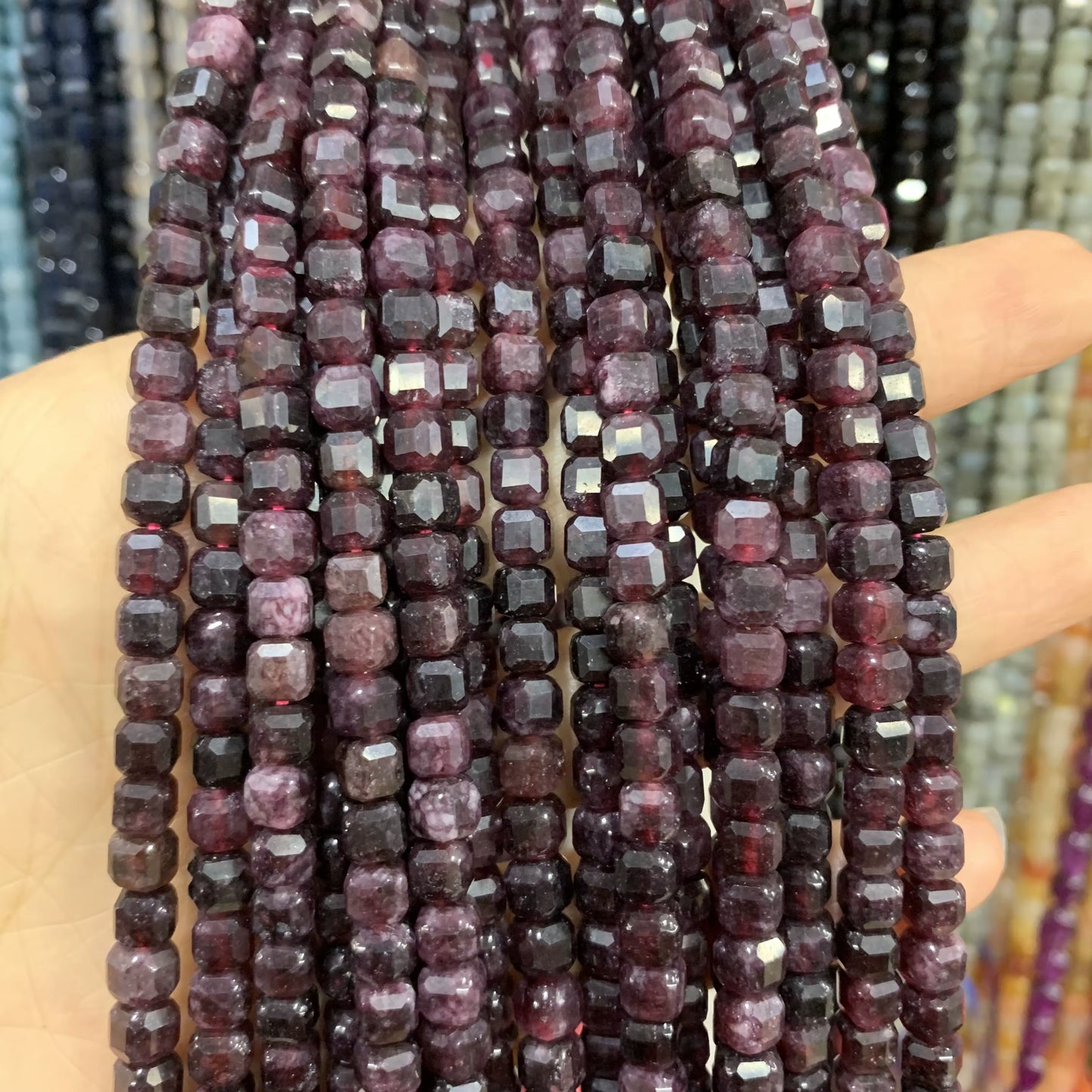 Faceted cube beads in dark purple and pinkish-red tones, strung on multiple strands and held in a hand against a blurred background of other beaded strands.