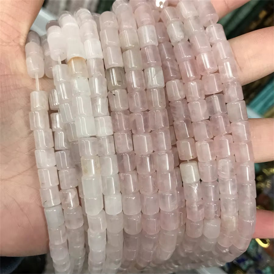 A hand holding multiple strands of cylindrical beads made from pale pink and milky white stone, showing the translucent texture and color variation.