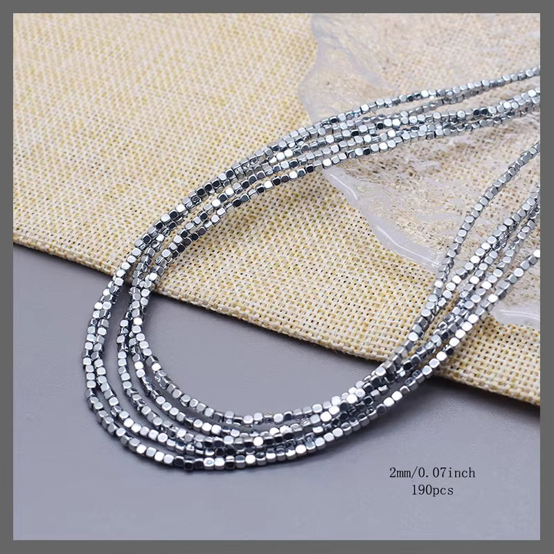 Five strands of silver faceted round beads, 2mm (0.07 inch) in length, displayed on a neutral gray surface with a beige woven fabric background.