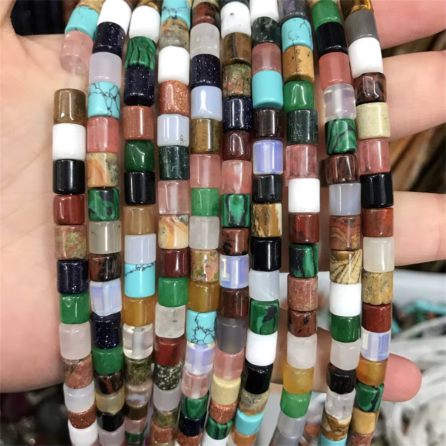 Multiple strands of cylindrical stone beads held in a hand, featuring a wide variety of colors and patterns including turquoise, green, brown, white, black, and marbled stones.