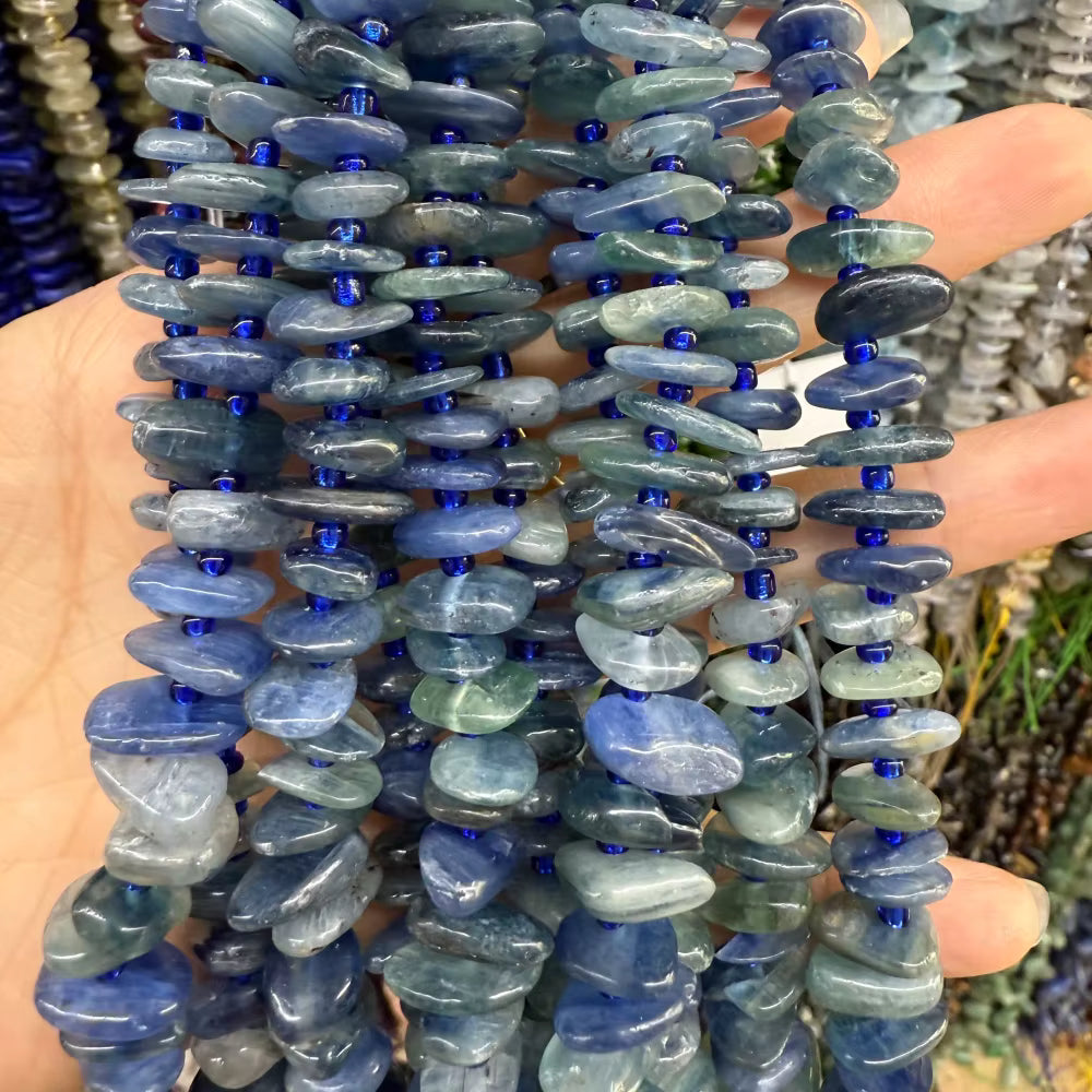 A close-up of multiple strands of polished blue tumbled stone beads, likely kyanite or sapphire, strung on dark cords and held in a hand. The beads are oval-shaped with a pearly, milky texture and a strong blue cast, interspersed with small dark blue seed beads.