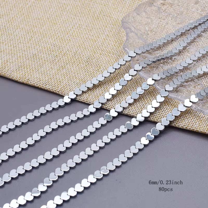 Five strands of silver heart shaped hematite beads  on a gray surface with a beige woven fabric backdrop.  text in the bottom right specifies 4x2mm/0.