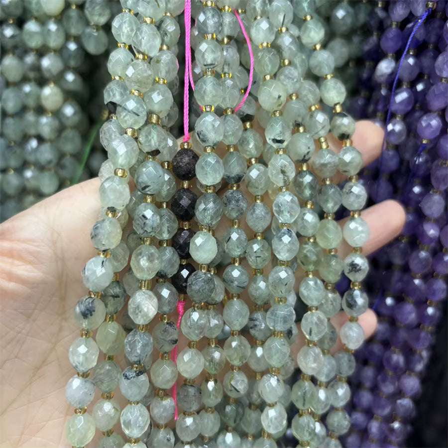 Faceted round beads strung on cords, held in a hand. The beads are primarily pale gray-green with black inclusions, separated by small gold spacer beads, with pink and purple cords visible.