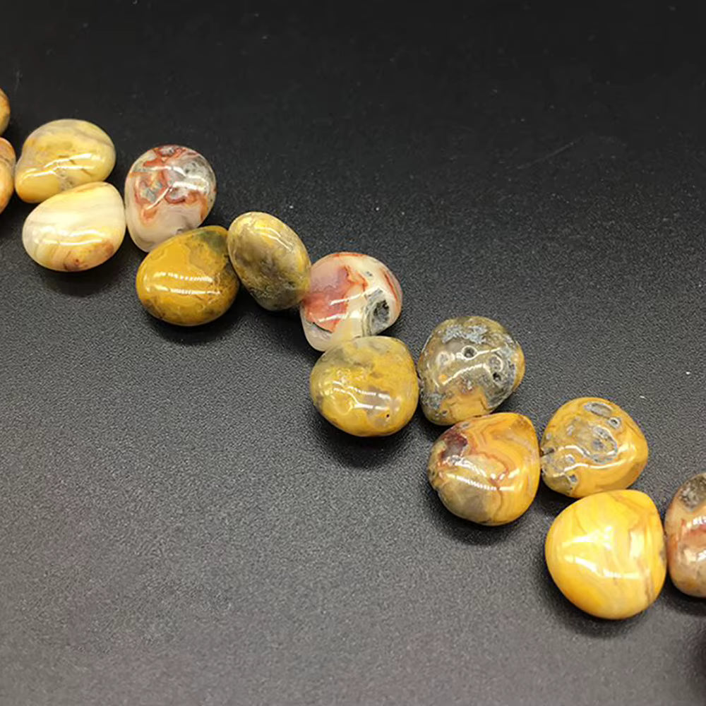A close-up of polished natural stone beads arranged diagonally on a dark gray background. The beads are smooth, rounded, and display natural marbled patterns in yellow, brown, cream, and gray.