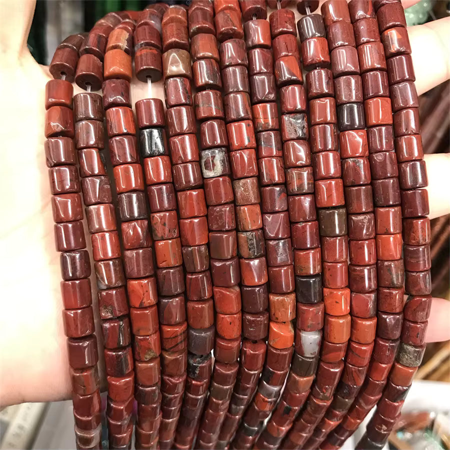 A hand holding a bundle of cylindrical red stone beads, likely jasper or agate, in multiple shades of red, brown, and marbled tones.