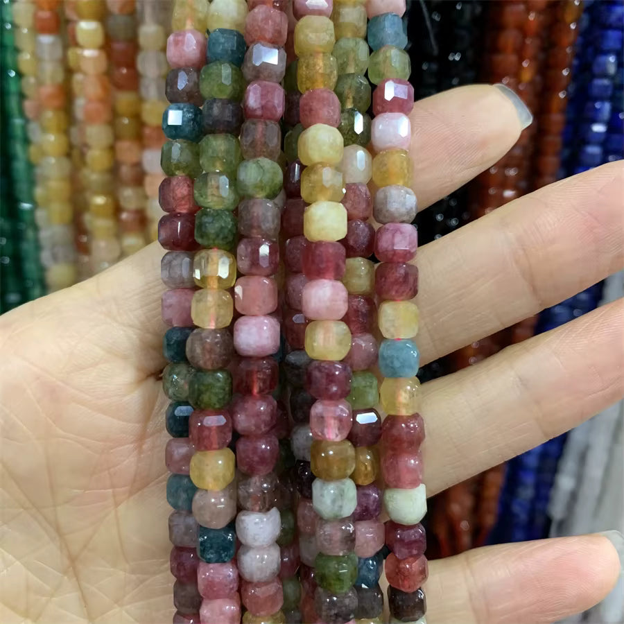 A hand holding multiple strands of faceted square beads in assorted pastel and earthy colors, with more colorful bead strands blurred in the background