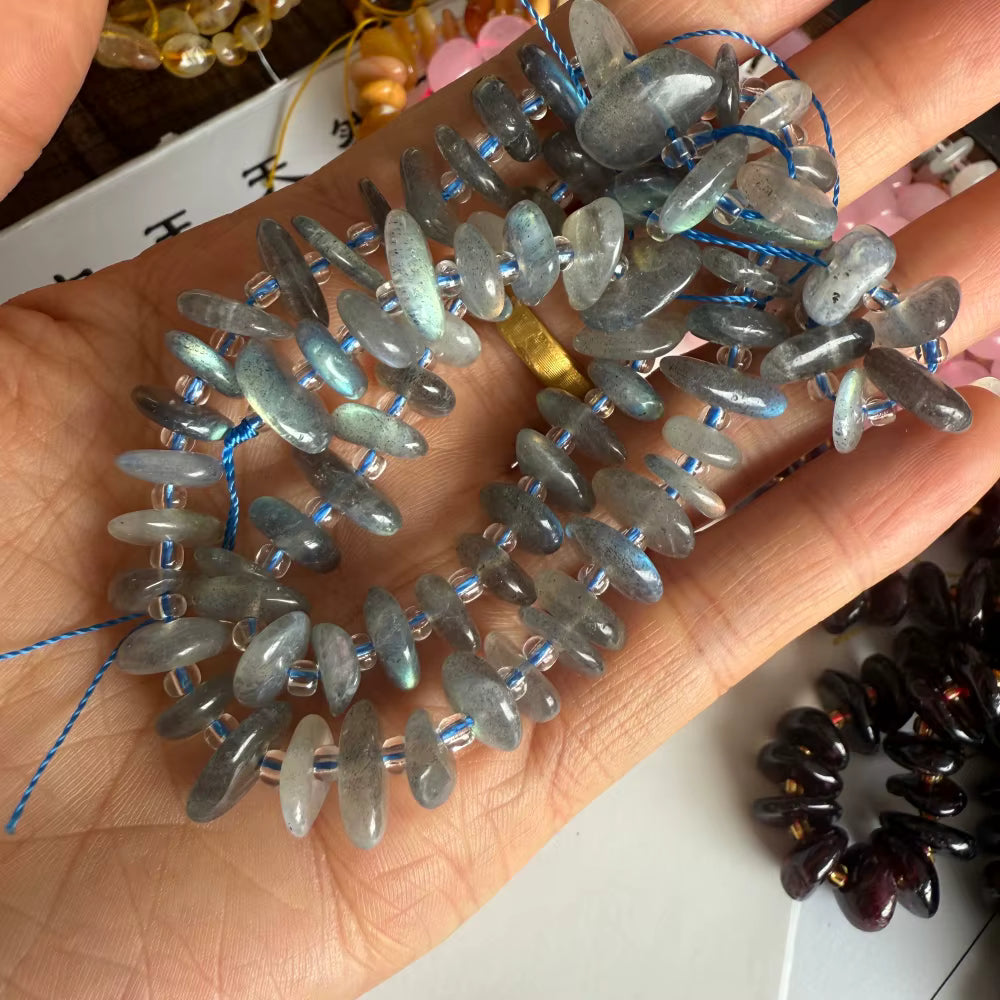 A hand holding a strand of gray iridescent stone beads threaded on blue string, with other beads partially visible in the background.