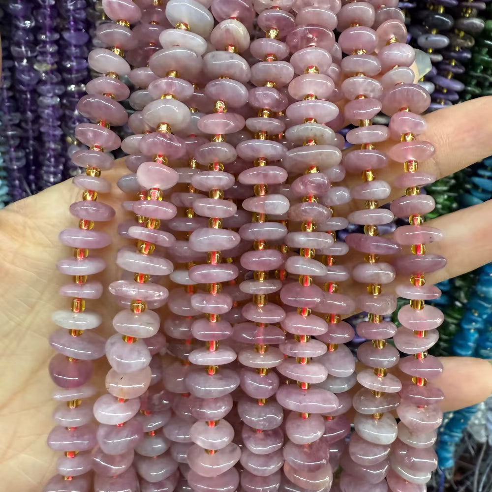 A close-up of a hand holding multiple strands of polished pink stone beads with small gold spacer beads, with other colorful bead strands blurred in the background.