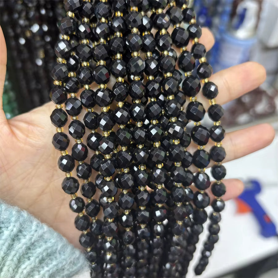 A hand holding multiple strands of faceted black beads with small gold spacer beads, shown in close-up against a blurred workshop background.