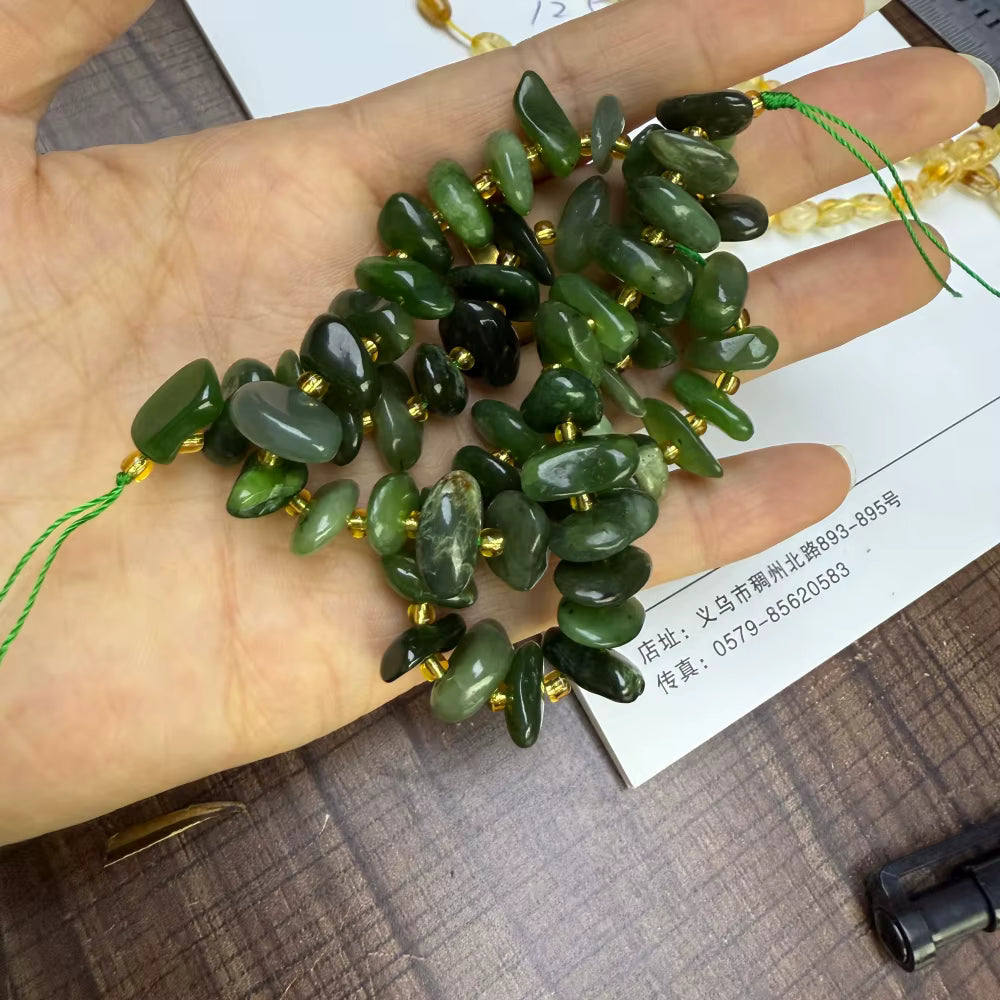 A hand holds a cluster of polished green stone beads strung on cords, with some dark green beads and small gold spacer accents. A white card with printed contact information lies underneath the beads on a dark wood surface.