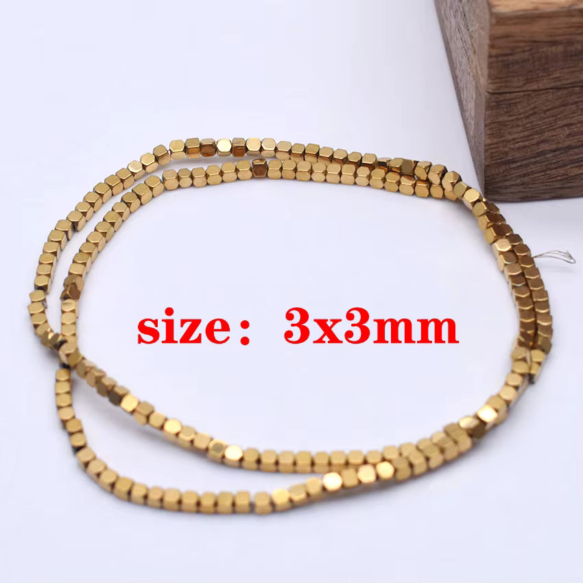 A coiled strand of small metallic gold rounded cube beads on a white surface, with red text reading 'size: 3x3mm' overlaid in the center and a wooden block partially visible in the top right corner.