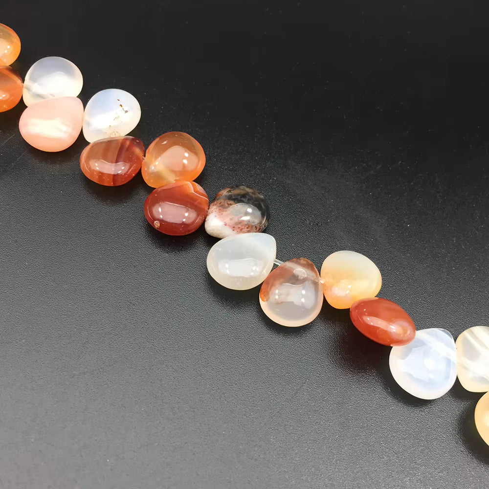 A close-up diagonal shot of polished agate stone beads in various natural colors including orange, red, white, and translucent cream, arranged on a dark textured surface.