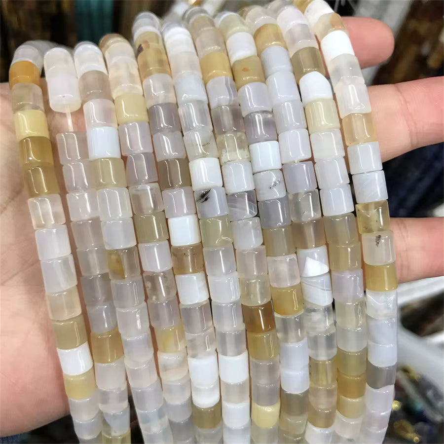 A hand holding multiple strands of cylindrical agate beads in natural stone colors including beige, white, gray, and light brown.