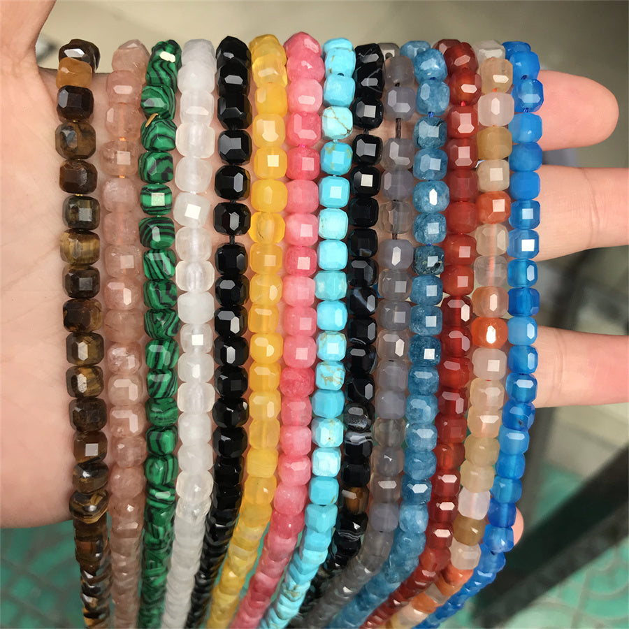 A hand holding multiple strands of faceted gemstone beads in various colors, including brown, pink, green, white, black, yellow, pink, turquoise, gray, red, and blue.