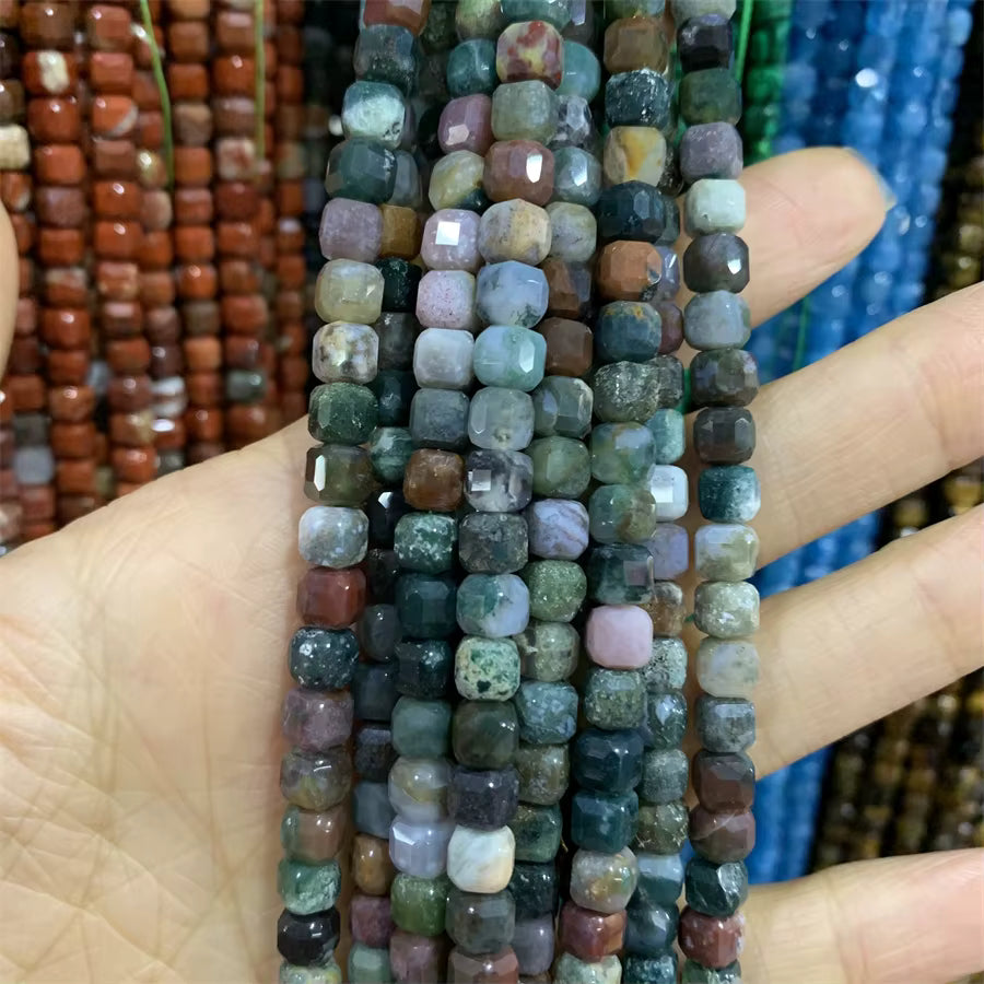A hand holding several strands of faceted gemstone beads in earthy, multicolored shades including green, brown, pink, gray, and white, with other colorful bead strands blurred in the background.
