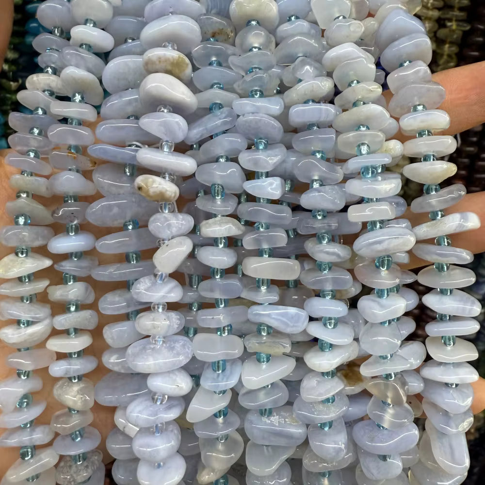 Multiple strands of polished tumbled stone beads in pale blue, lavender, gray, and white tones, strung on clear cords with small metallic spacer beads, held in hands against a dark background.