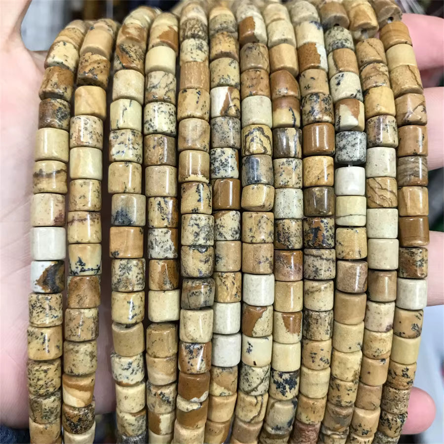 A close-up of multiple strands of cylindrical stone beads strung together, held in a person's hand. The beads are made of natural stone with varied colors including tan, brown, cream, and dark gray veining.