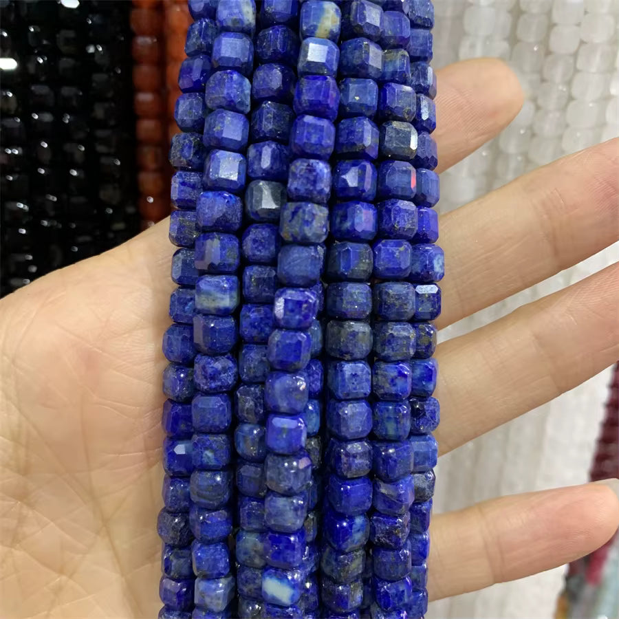 A hand holding several strands of faceted cobalt blue lapis lazuli cube beads, with other colored bead strands blurred in the background.