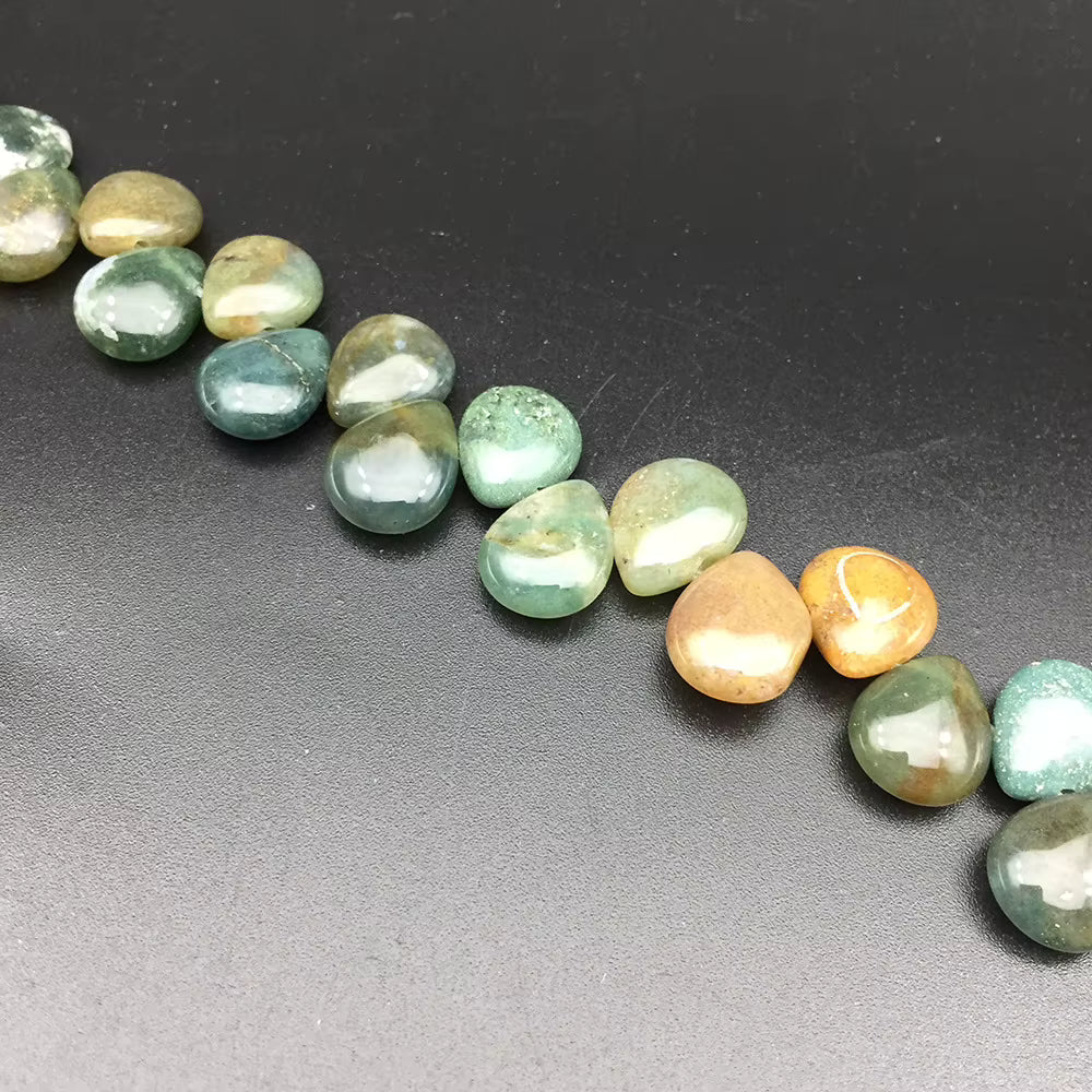 A diagonal strand of polished tumbled gemstone beads in shades of green, blue-green, and orange, displayed on a dark gray textured background.
