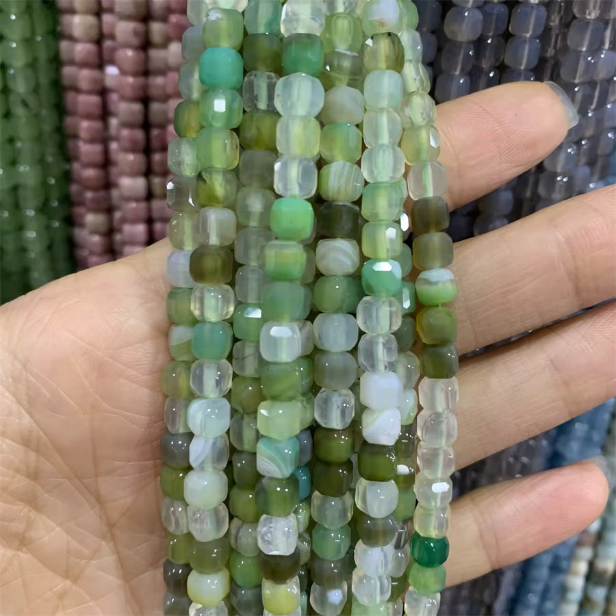 Multiple strands of faceted cube beads in various shades of green, olive, and translucent white, held in a person's hand.