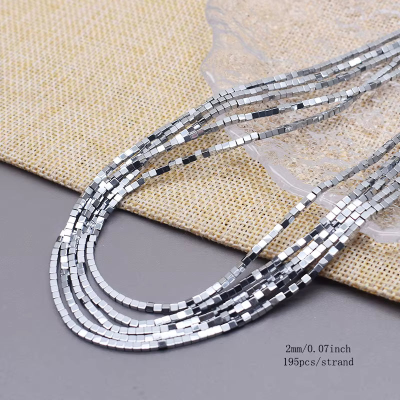 Five strands of silver cube hematite beads  on a gray surface with a beige woven fabric backdrop. text in the bottom right specifies 2mm/0.