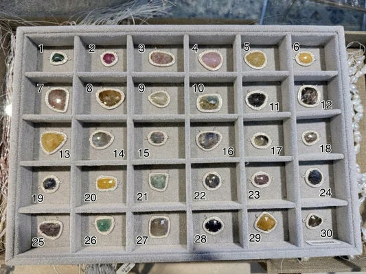 A gray ring display tray with 30 numbered compartments, each holding a different gemstone ring, photographed from above.
