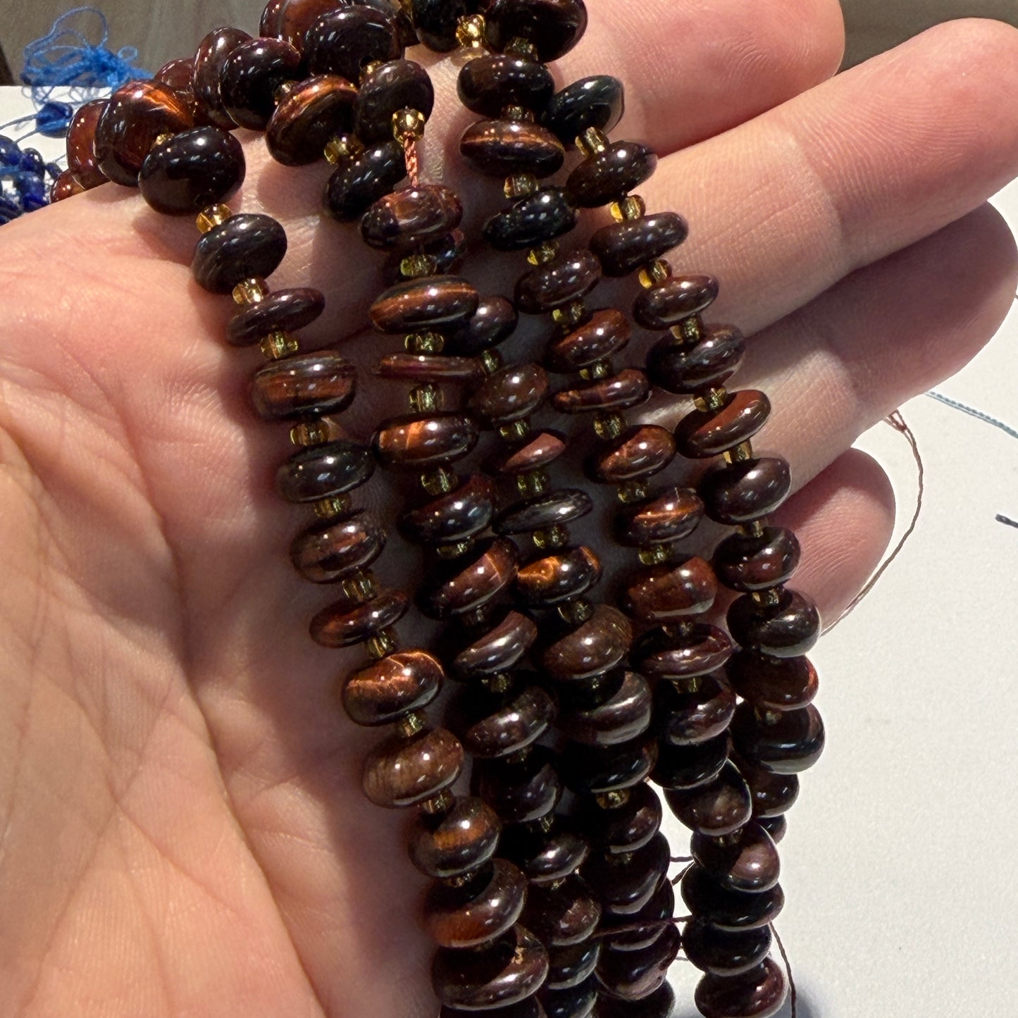 A close-up of a hand holding several strands of polished dark brown and black stone beads with warm golden-brown highlights, separated by small metallic spacer beads.