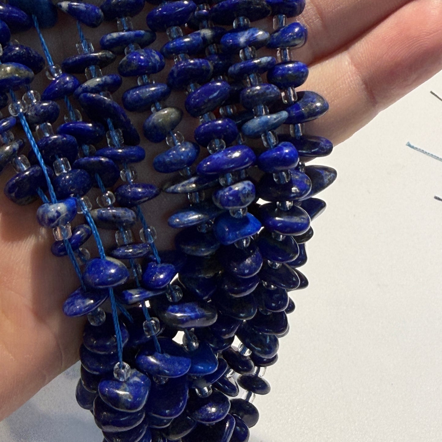A close-up of a hand holding a strand of polished lapis lazuli crystal beads on blue cord, with multiple strands layered together against a light background.