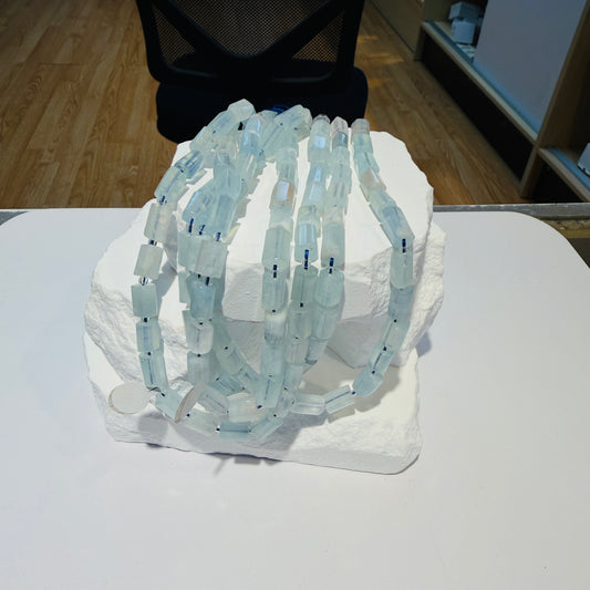 A set of translucent pale blue faceted bead strands arranged on a white display block, photographed indoors on a white table with a black chair and wood floor in the background.