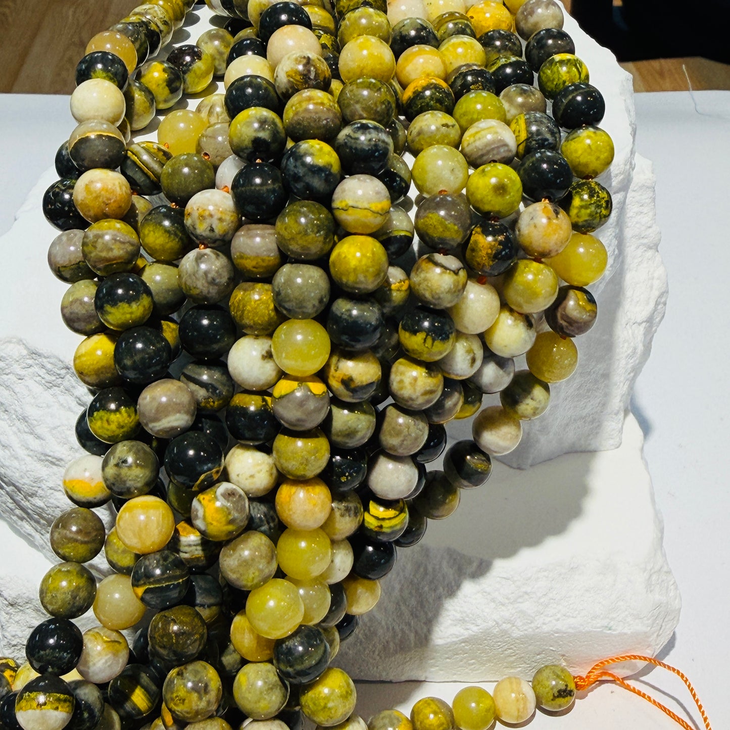 Multiple strands of polished round beads draped over a white stone display block. The beads are marbled with black, olive green, yellow, and cream tones, with a small orange cord visible at the bottom right.