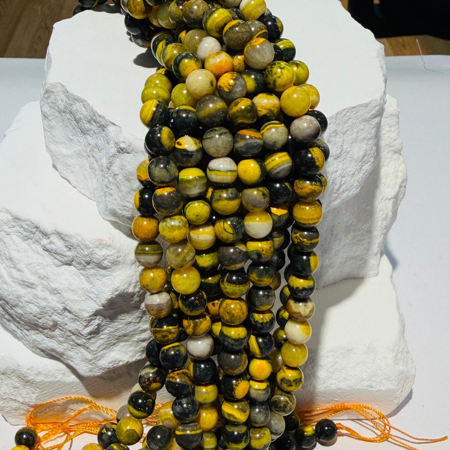 Multiple strands of polished round beads draped over white display stones. The beads are primarily yellow and black with some gray, white, and brown marbled variation.