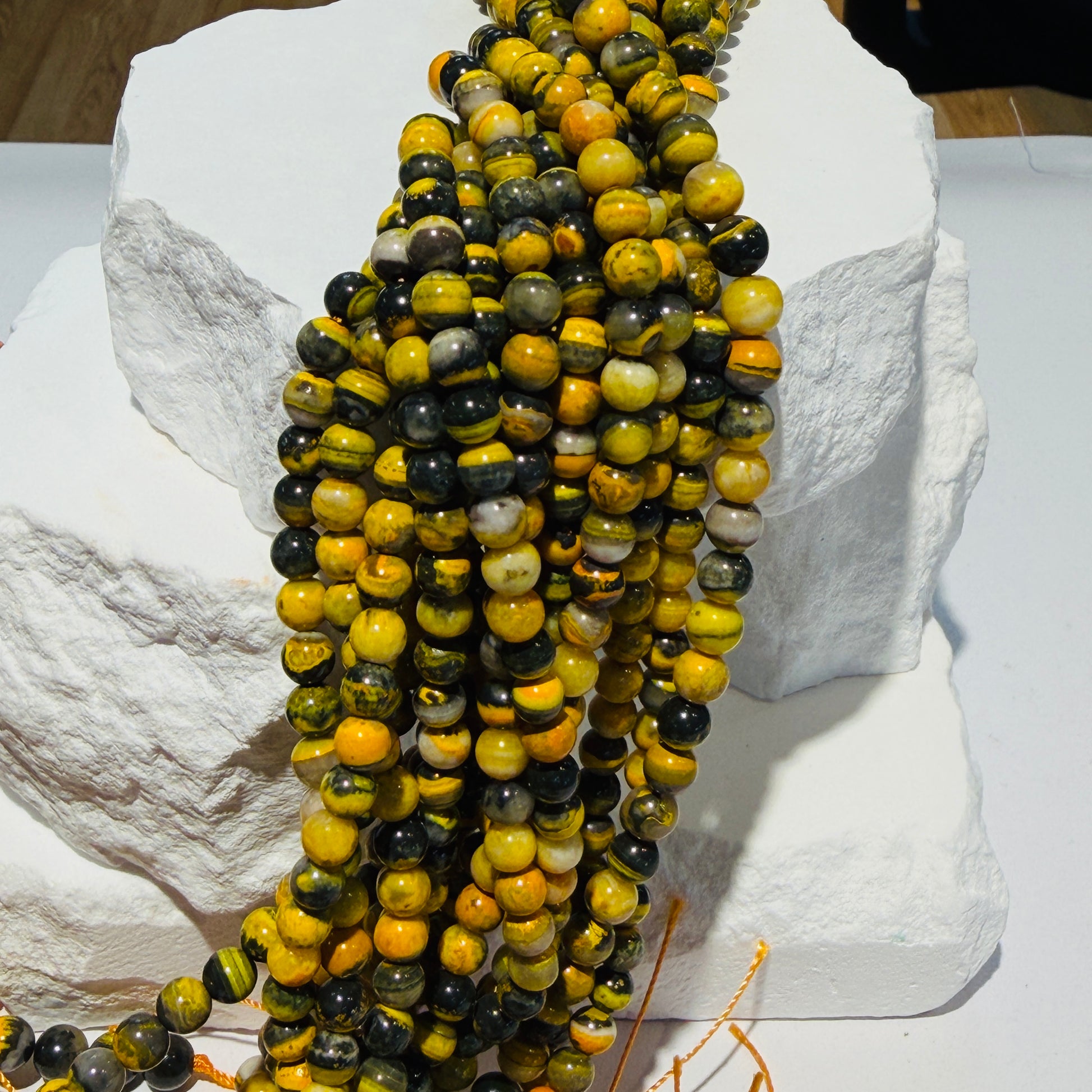 Multiple strands of polished round beads draped over white stone blocks. The beads are marbled with yellow, black, green, and orange tones.