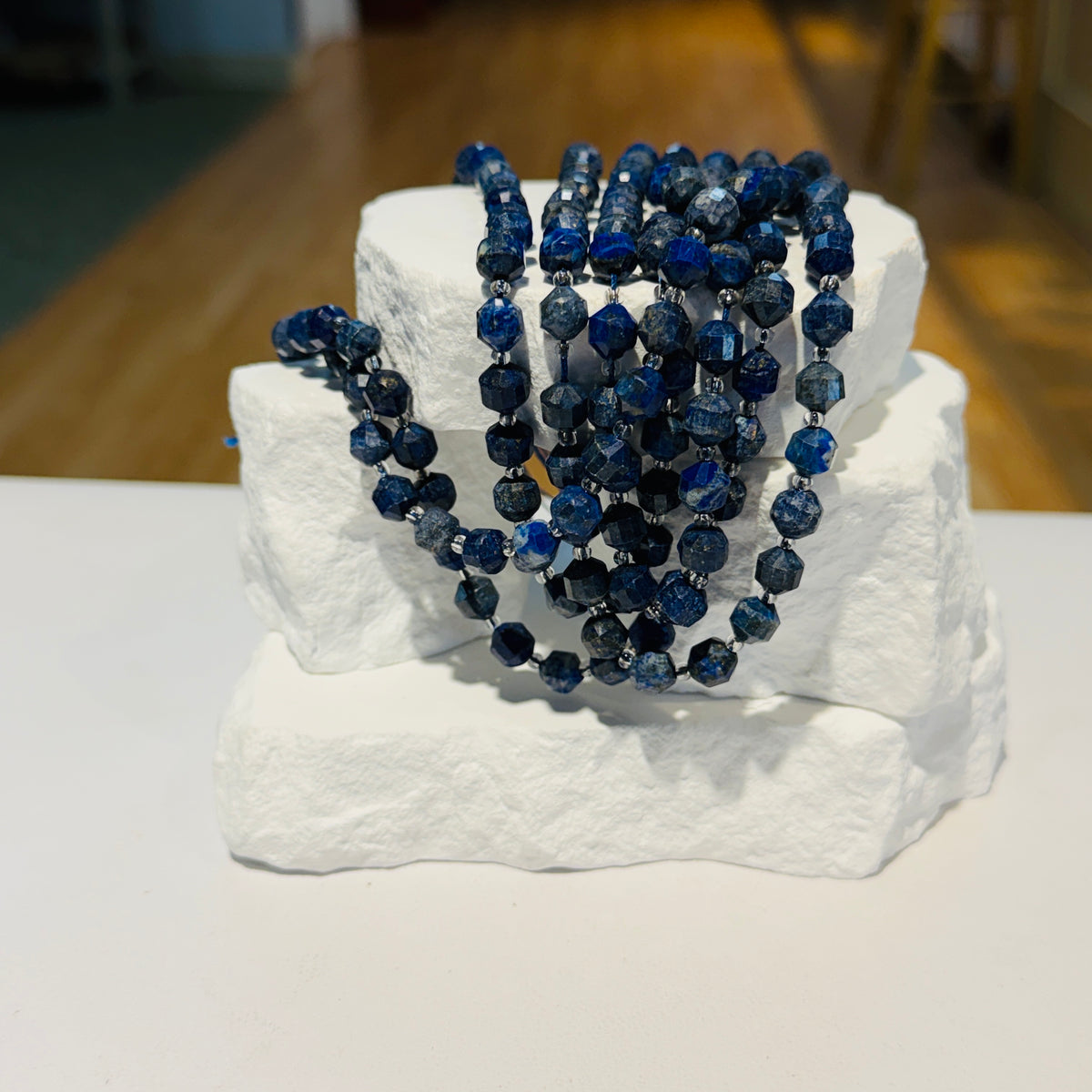 strands of faceted Lapis beads draped over stacked white display blocks on a white tabletop, with a softly blurred indoor background.