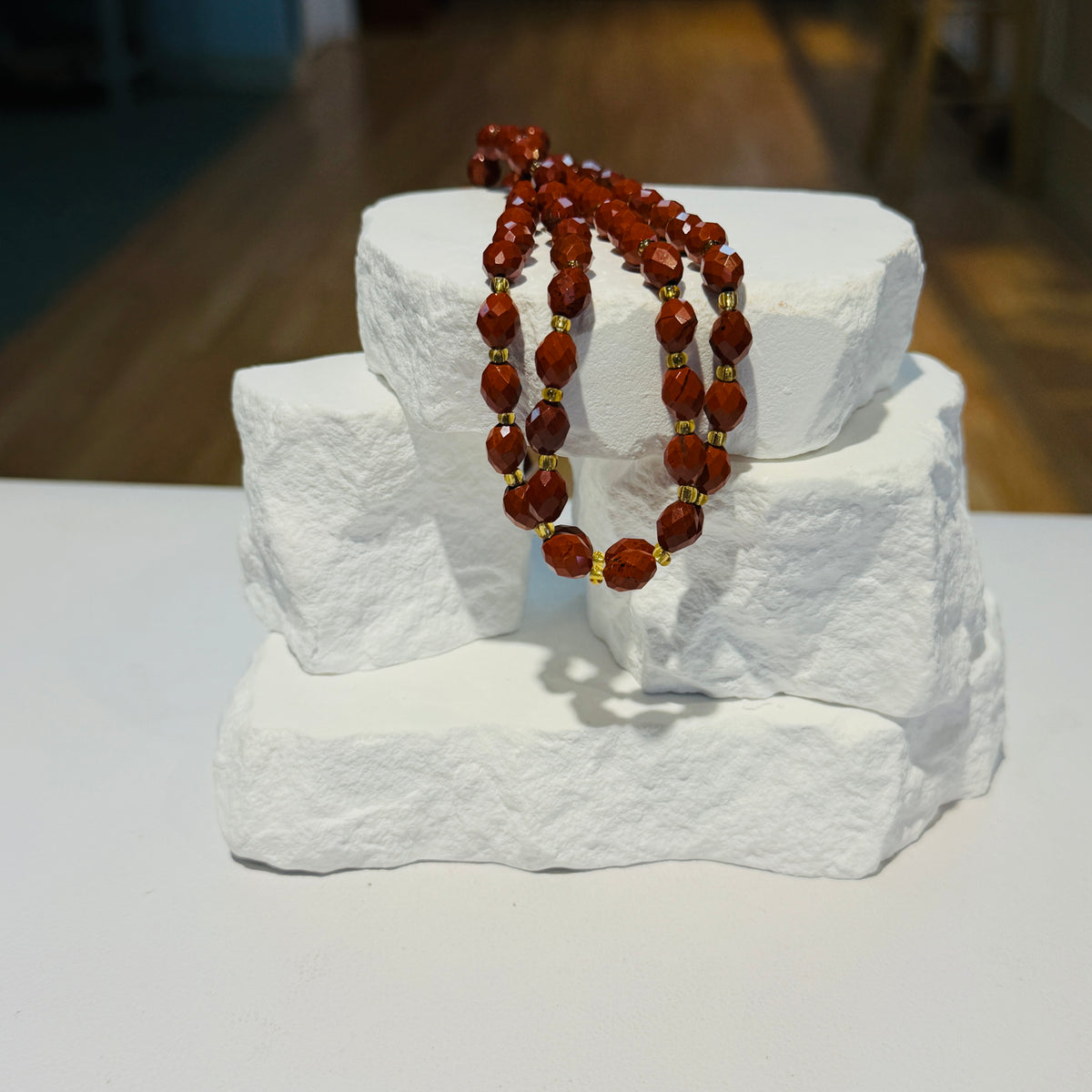 A strand of Red Jasper  Beads separated by small gold spacer beads, draped over a white stone display block on a white surface.